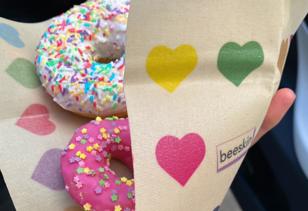 2 donuts, one with a pink, one with a white glazing and colorful sprinkles on top of it.  All is wrapped in a beeskin with colorful hearts. we can see the hearts in the front in pink, yellow and green next to a beeskin logo.