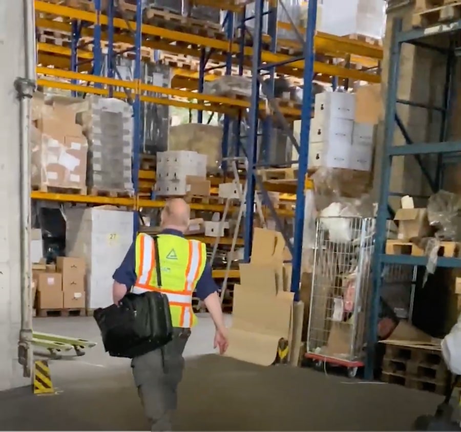 picture shows inspection officer entering a high-bay warehouse. he wears a neon yellow vest with TÜV Logo on it. 