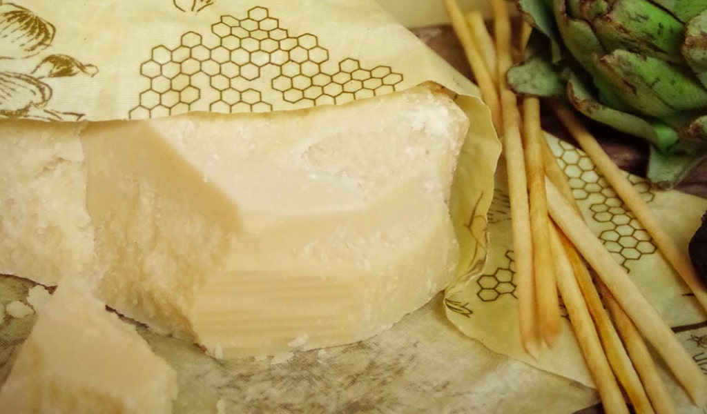 showing a big chunk of parmigiano reggiano wrapped in beeskin. showing also some grisini bread sticks and an artichoke at the top right of the image.