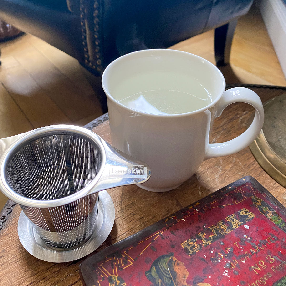 white porcelaine cup filled with hot water next to tea infuser with beeskin logo standing on a table