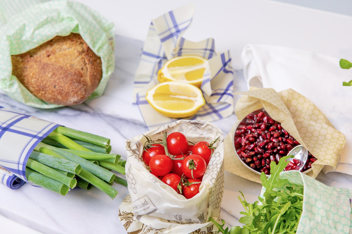 Image showing different food wrapped in beeskin. beeskin bag small in flower design holds tomatoes, beeskin bag L in standard design with honeycombs shows bread. Lemon and leek onions wrapped in Beeskin Beeswax wraps in kitchen towel design