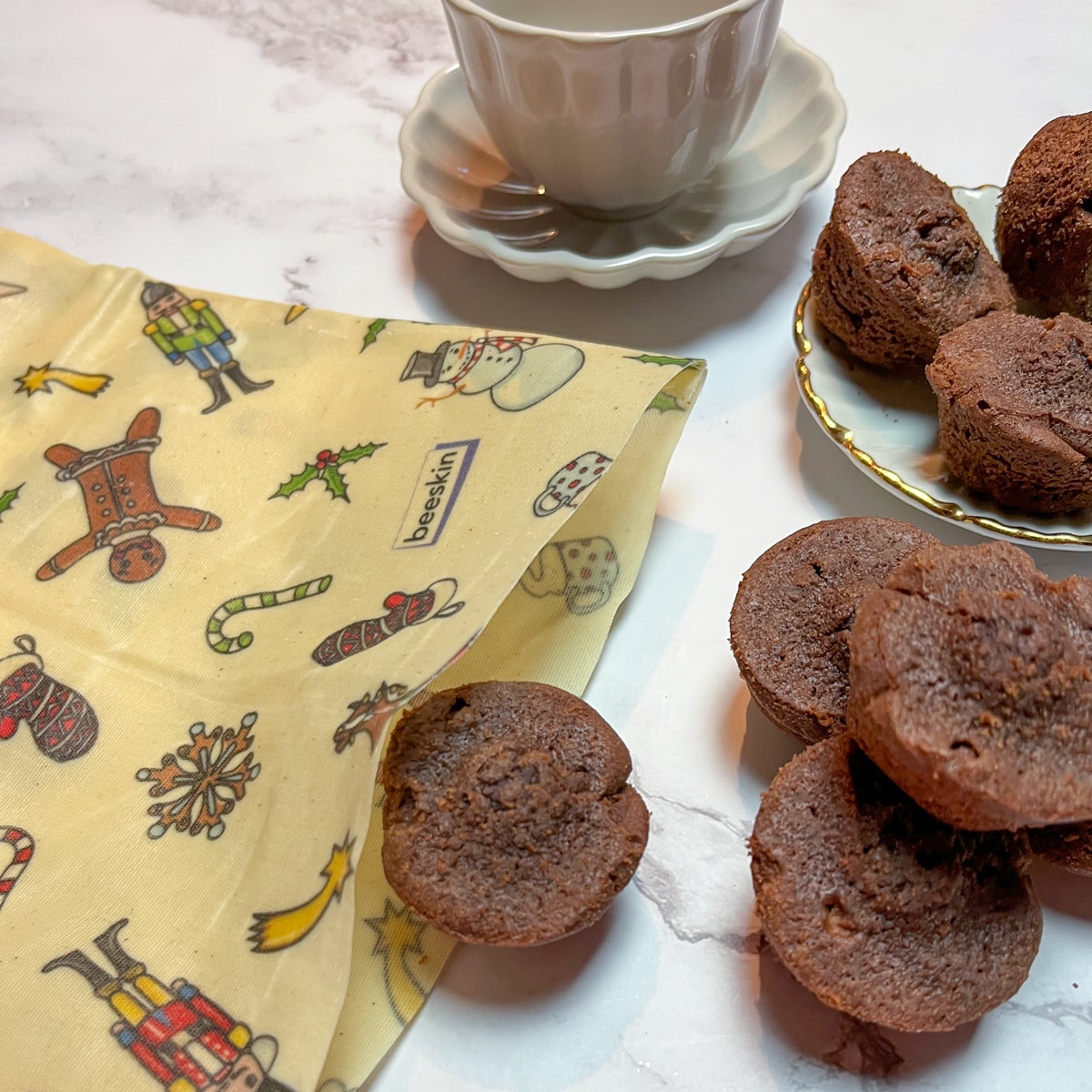 showing a beeskin beeswax bag in Christmas design with small images such as nutcrackers or stars. To the right are little brownies and at the back of the image is a small cup of coffee. 