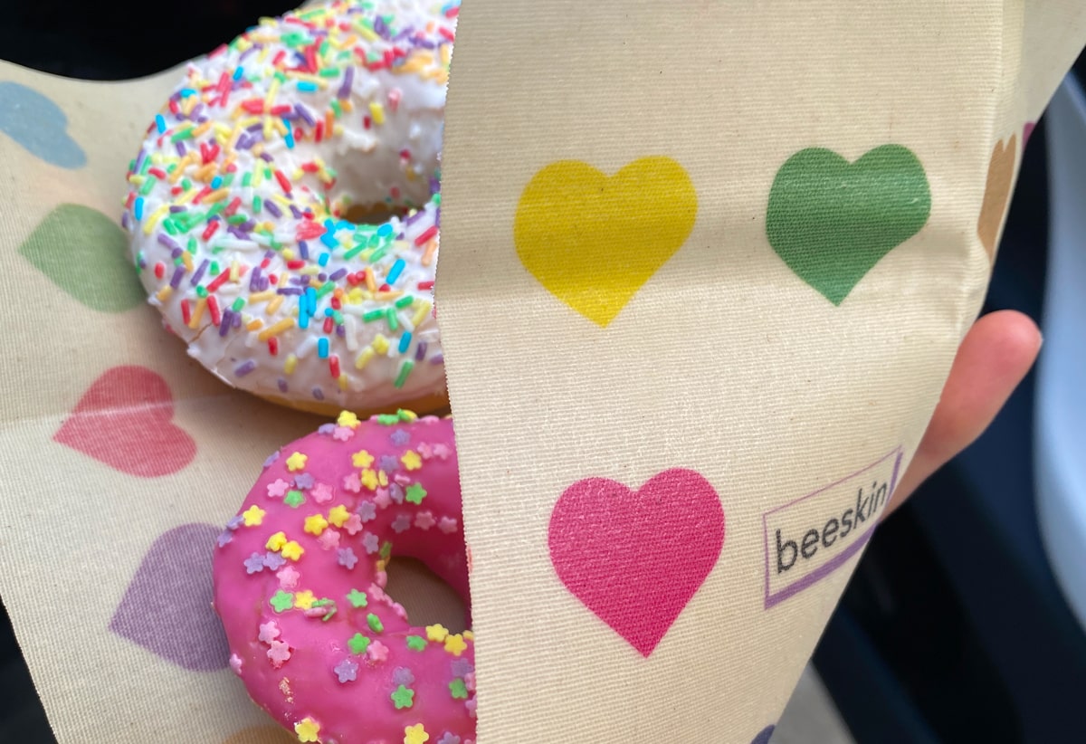 2 donuts, one with a pink, one with a white glazing and colorful sprinkles on top of it.  All is wrapped in a beeskin with colorful hearts. we can see the hearts in the front in pink, yellow and green next to a beeskin logo. 