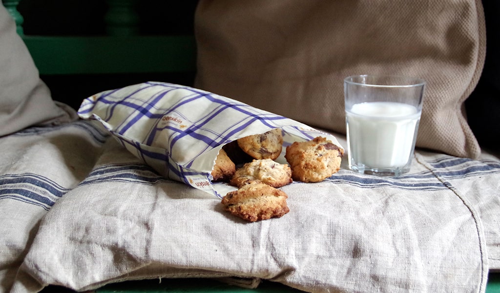 a bag full of cookies in a beeskin beeswax wrap bag in kitchen towel design. shown on linen covers and a glass of milk on the side. 
