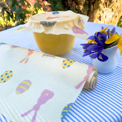 yellow bowl covered with beeskin easter edition and easter beeswax roll on a table with blue striped tablecloth a d blue/yellow flowers