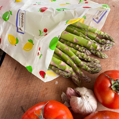 green Asparagus lies in a beeskin beeswax bag printed with fruits on a wooden table surrounded by garlic and tomatoes